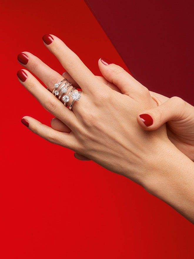 Hand mit rot lackierten Nägeln vor rotem Hintergrund trägt verschiedene Solitaire-Ringe mit "wachsenden Diamanten" am Mittelfinger