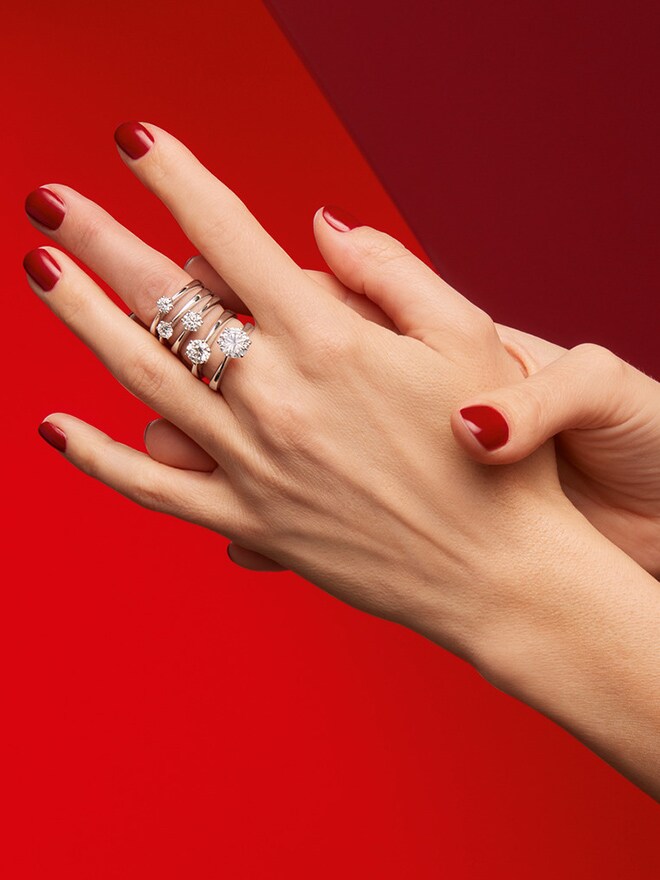 Eine Hand mit verschieden Solitaire-Ringen der Line One Renaissance by Wempe Classics vor rotem Hintergrund.