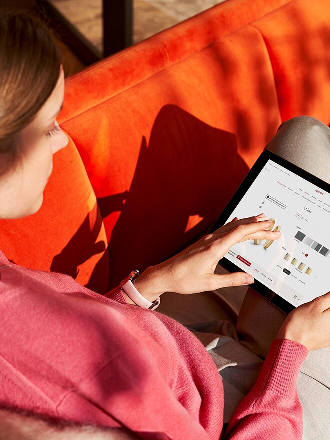 Frau sitzt auf orangefarbenem Sofa und betrachtet den Ehering-Konfigurator auf einem Tablet