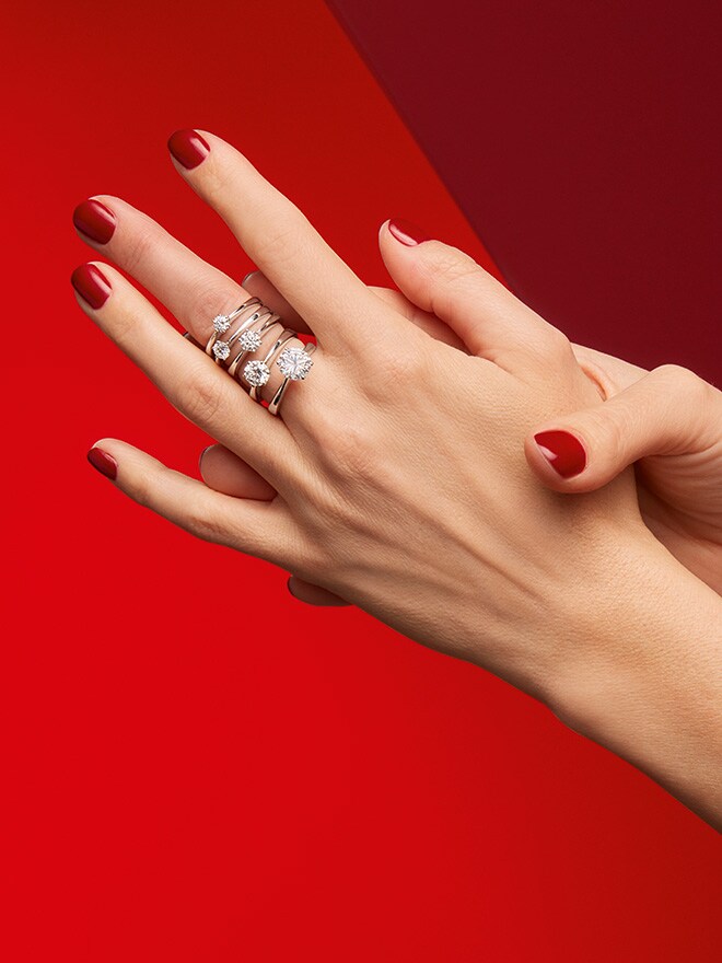 Zwei aneinander gelegte Frauenhände mit rotem Nagellack vor rotem Hintergrund. An einem Mittelfinger sitzen mehrere weißgoldene Solitaire-Ringe der Linie One 
