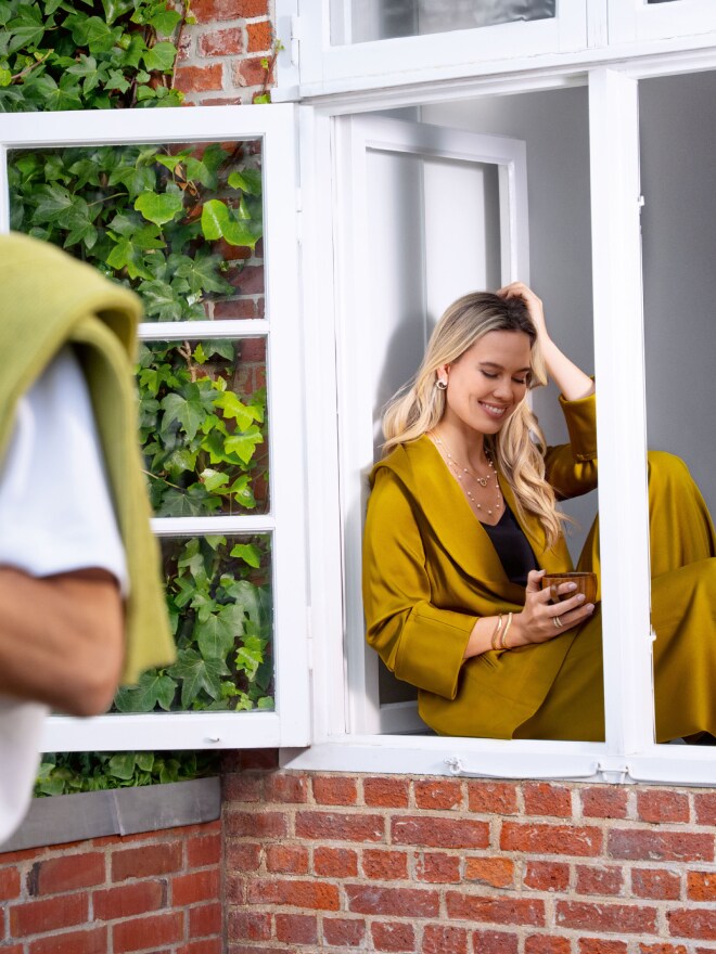 Blonde woman in an ochre-colored pantsuit sitting in an open window